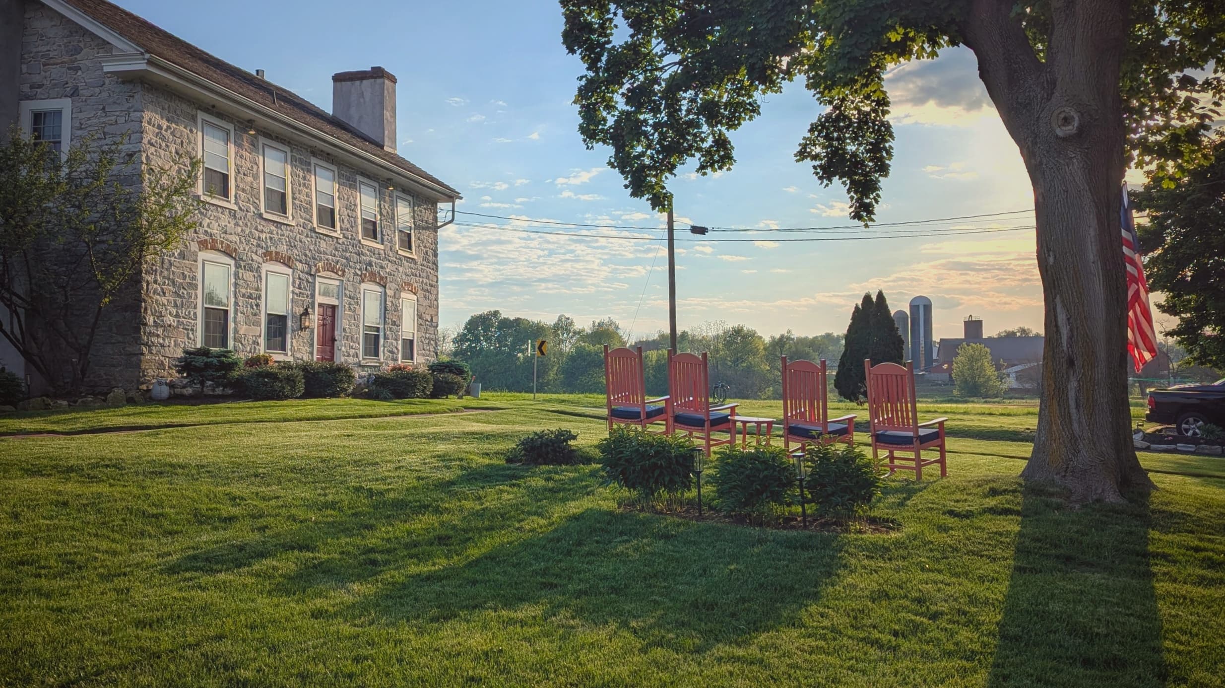 A sunny lawn features a stone house and a set of red chairs under a tree.