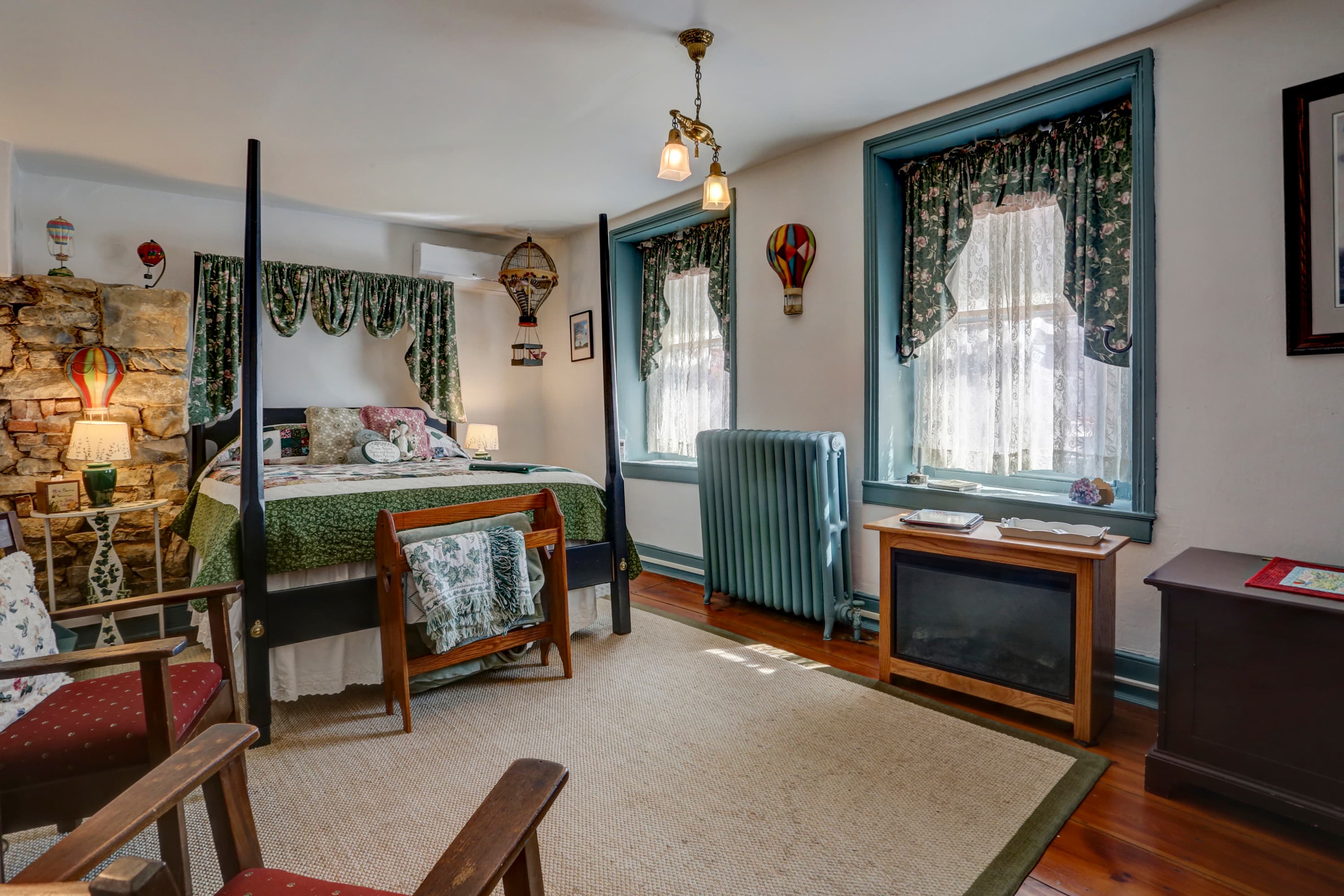 A cozy bedroom with a dark wood four-poster bed, two armchairs with red cushions, a small fireplace, and two windows with blue trim and floral curtains.