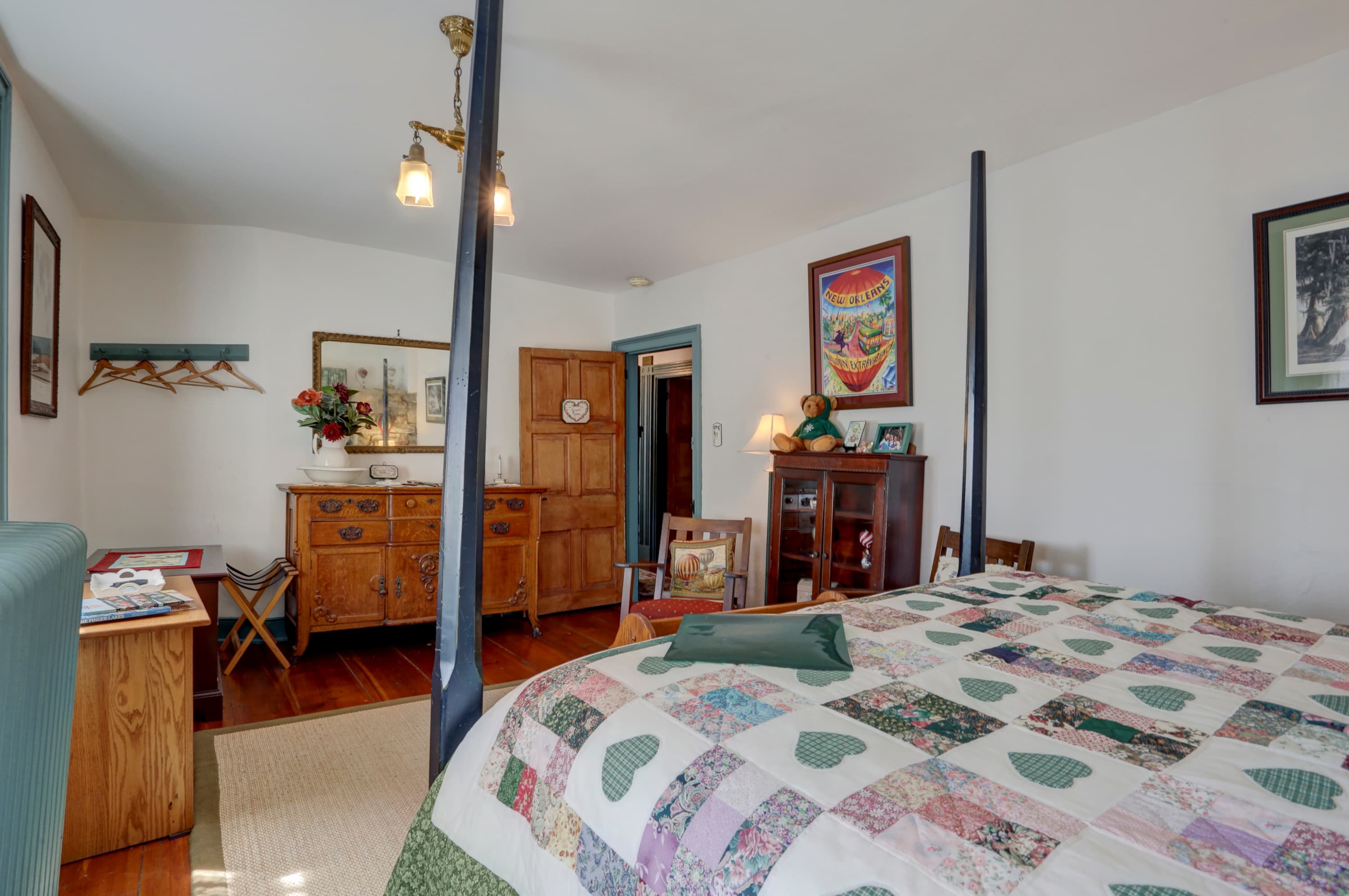 Bedroom featuring a four-poster bed with a patterned quilt, a wooden dresser with a mirror and flowers, and framed artwork.