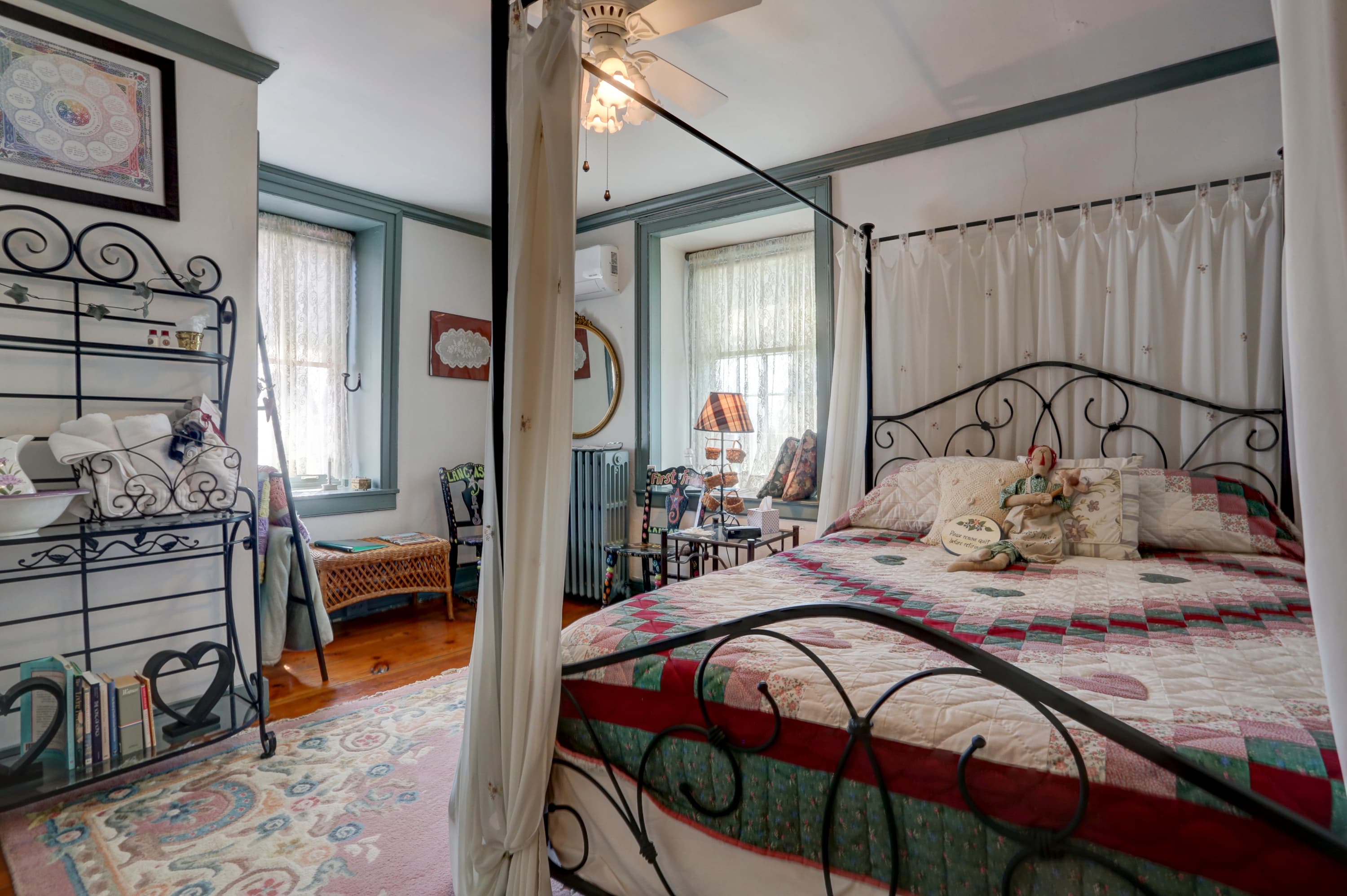 A bright and airy bedroom with a decorative iron bed with a patterned quilt, two windows with white curtains, a dresser with a mirror, and a patterned rug on the floor.