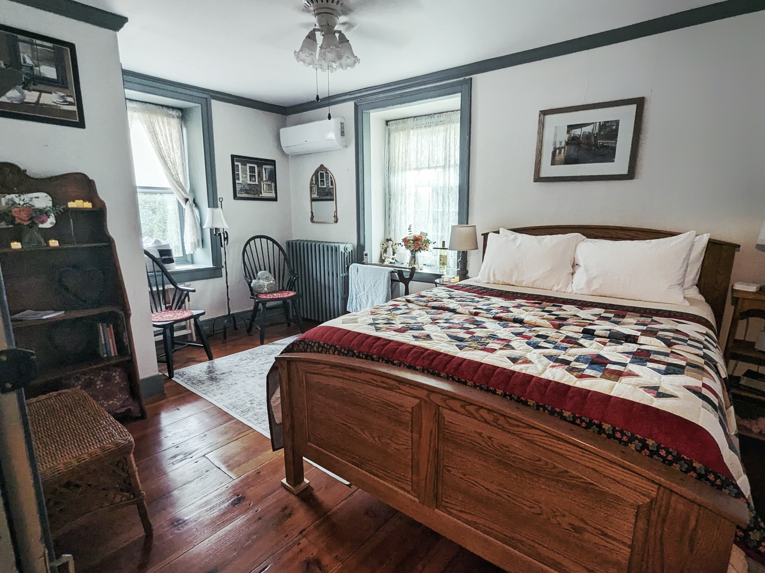 A bright and airy bedroom with a cottage style bed with a patterned quilt, two windows with white curtains, a dresser with a mirror, and a patterned rug on the floor.