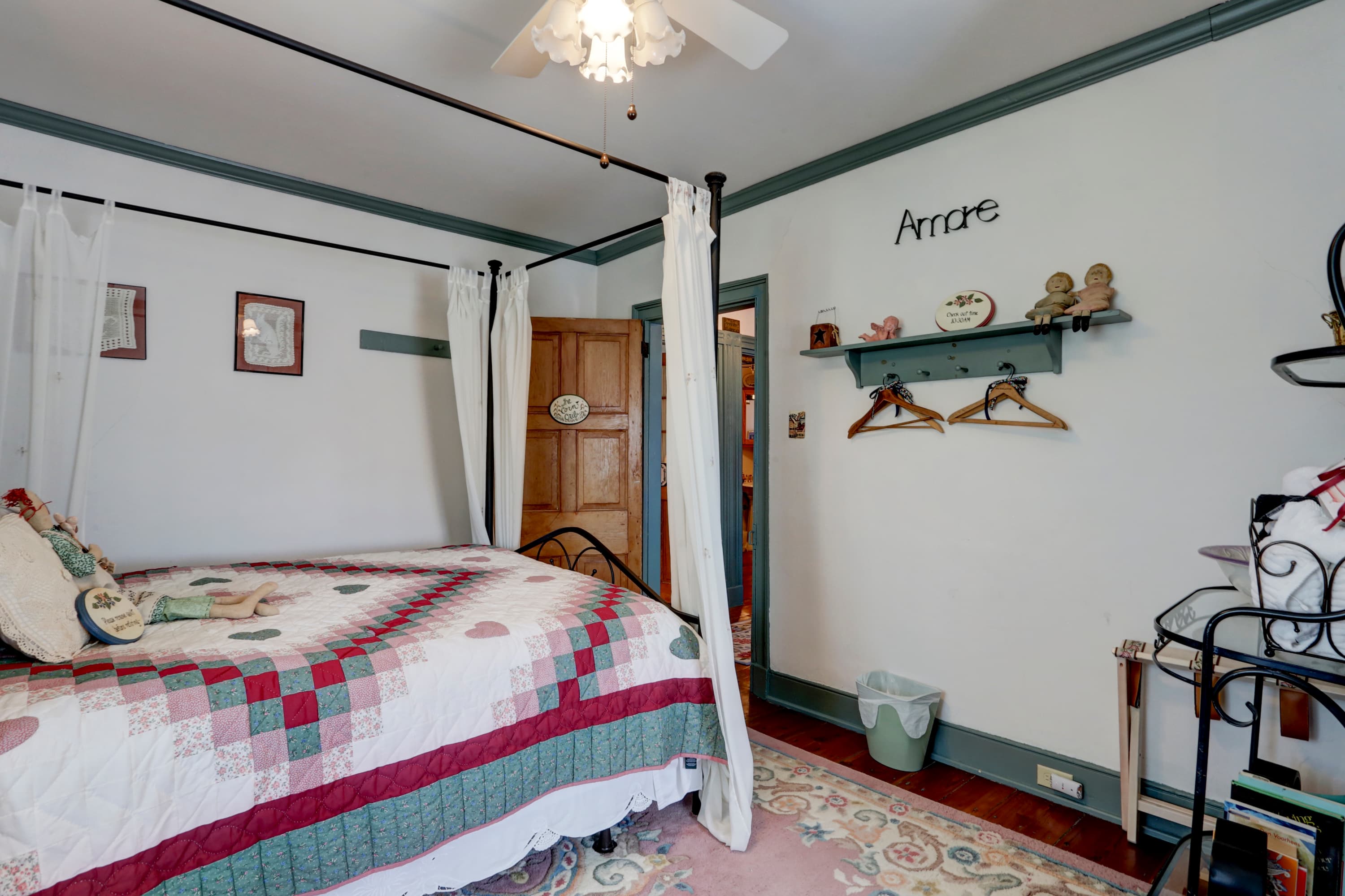 A bedroom with a white four-poster bed with a patterned quilt, a doorway leading to another room, and a wall shelf with decorative items.