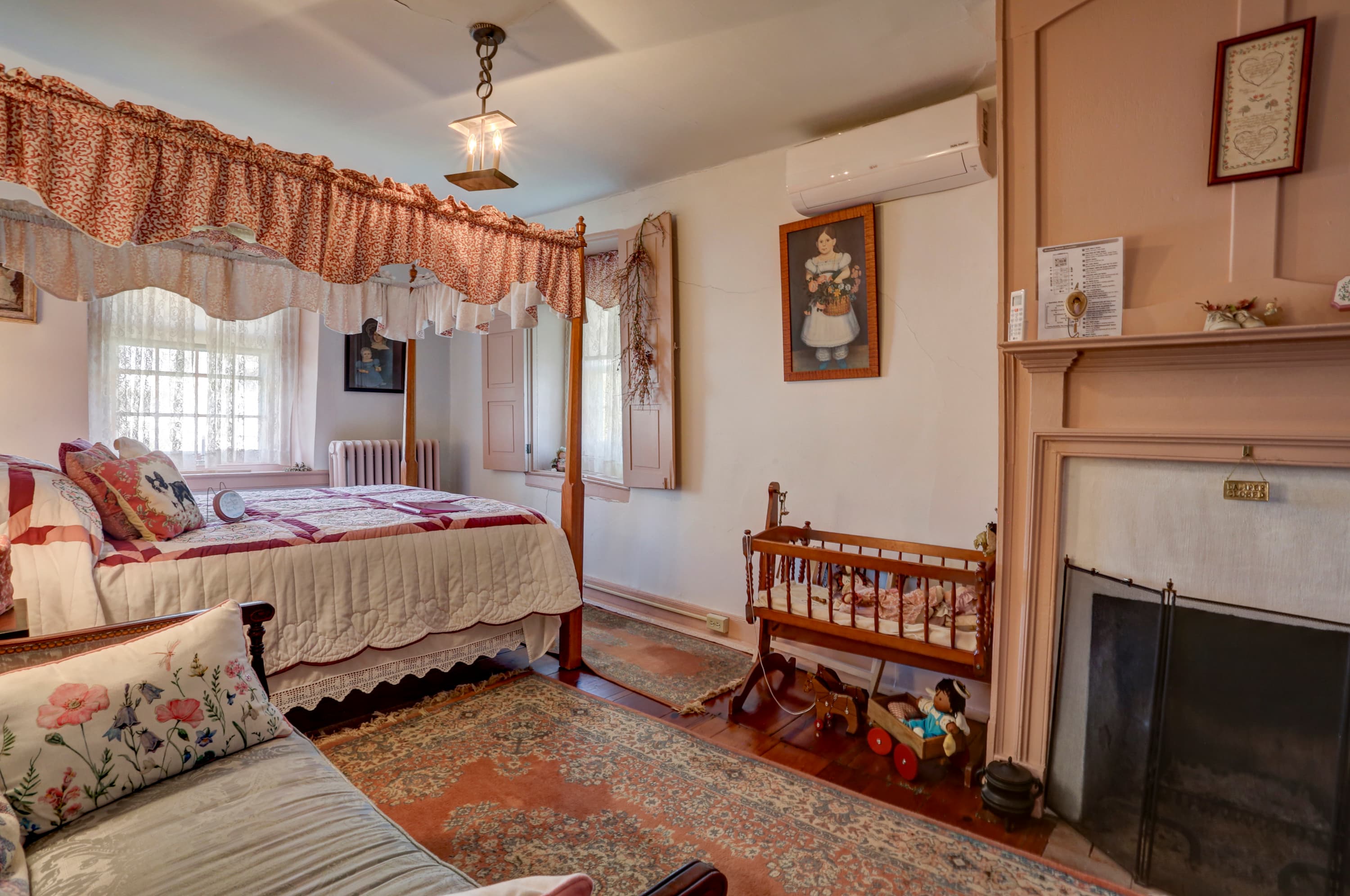 A cozy bedroom with a wooden canopy bed with a white and pink comforter, a fireplace with a pink mantel, a small wooden crib, and a patterned rug on the floor.