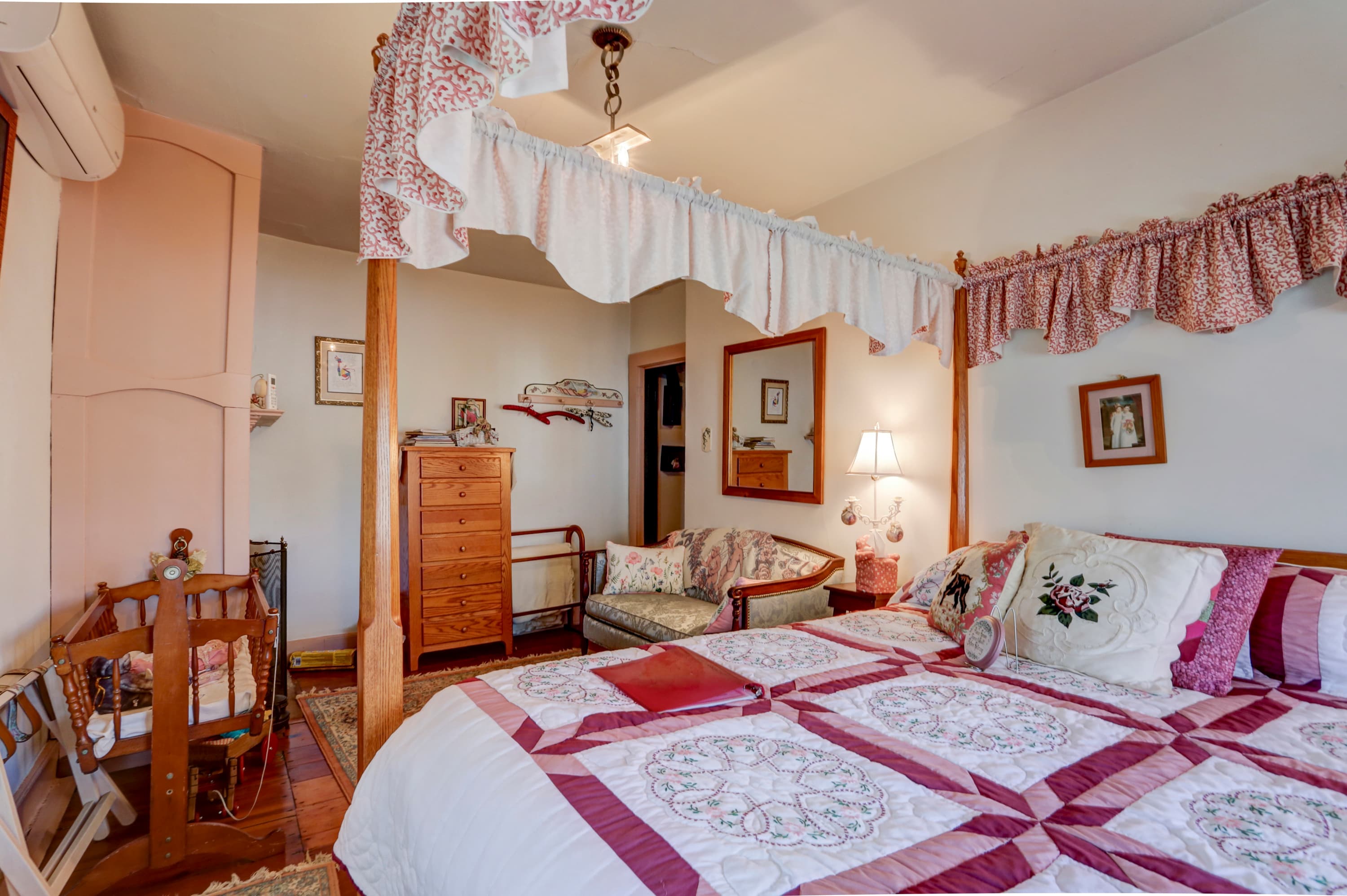 A cozy bedroom with a wooden canopy bed with a white and pink comforter, a wooden crib, a wooden dresser, and a patterned rug on the floor.