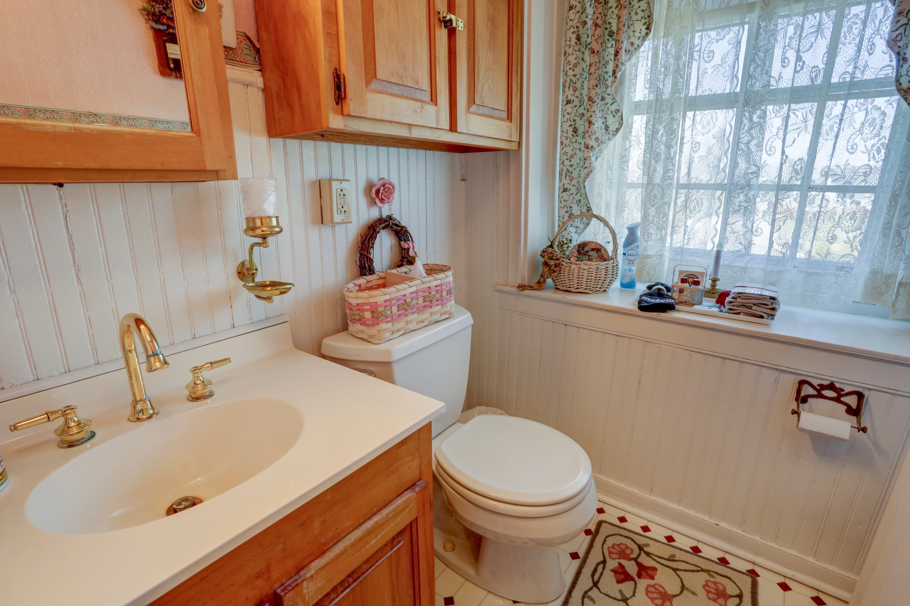 A bright and airy bathroom with a sink, a toilet, a window with a curtain, and a patterned rug on the floor.