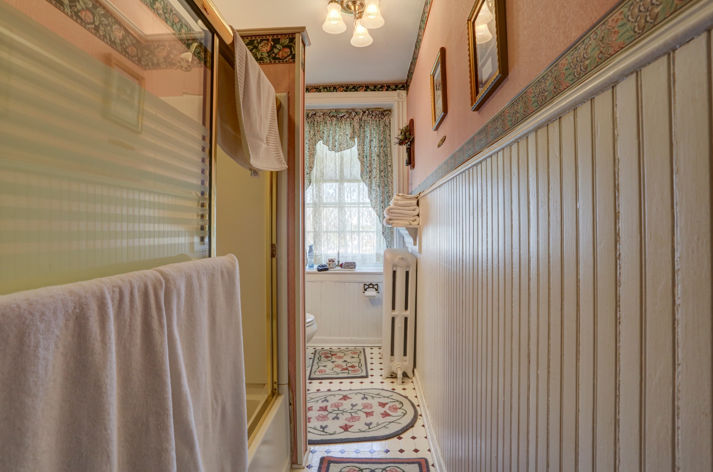 A hallway with pink walls, white wainscoting, and a patterned border at the top of the walls, leading to a small bathroom with a shower, toilet, and patterned rug on the floor.