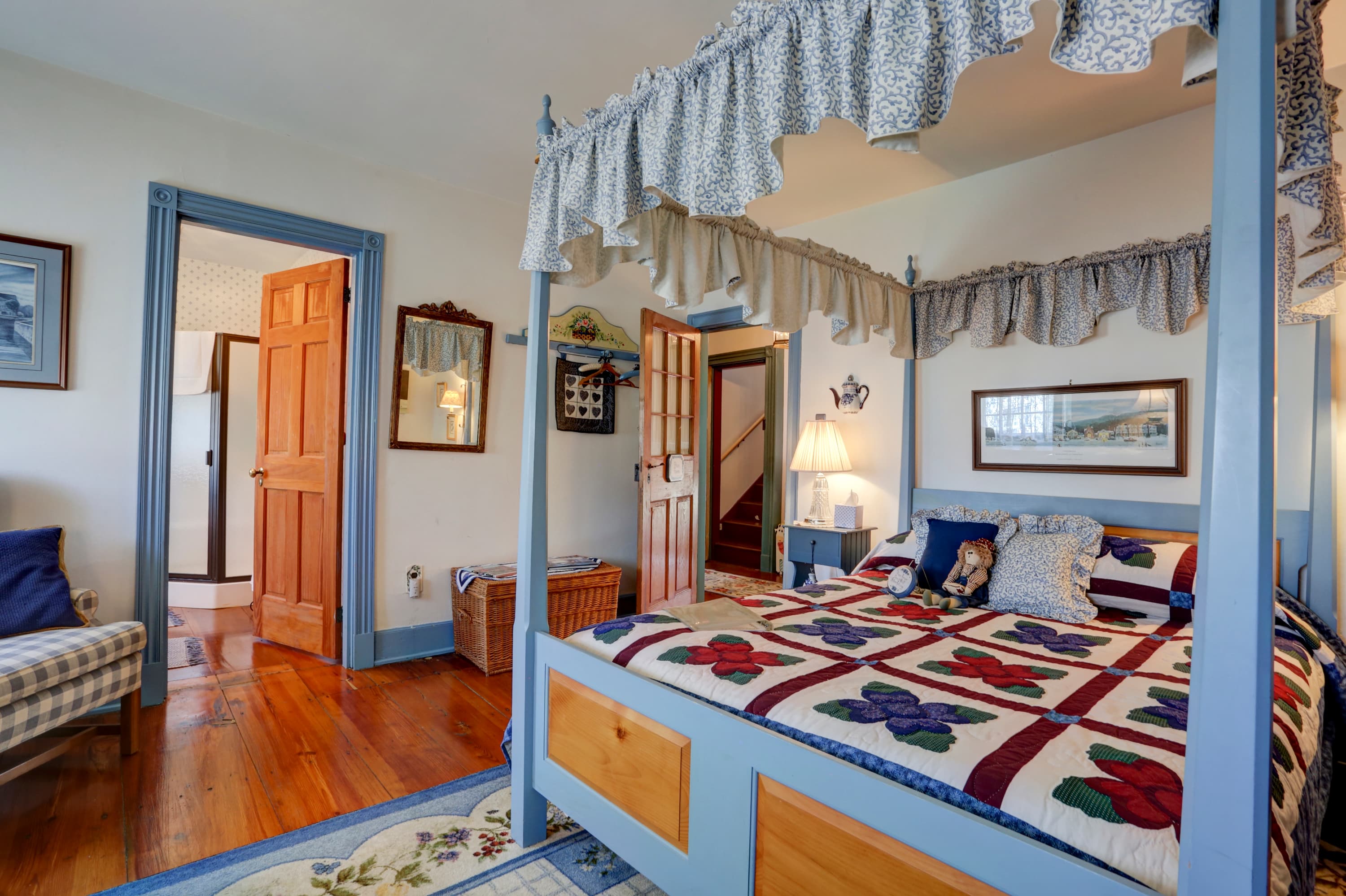 A charming bedroom with a blue and white four-poster bed with a white canopy, a doorway leading to another room, a wooden dresser, and a patterned rug on the floor.