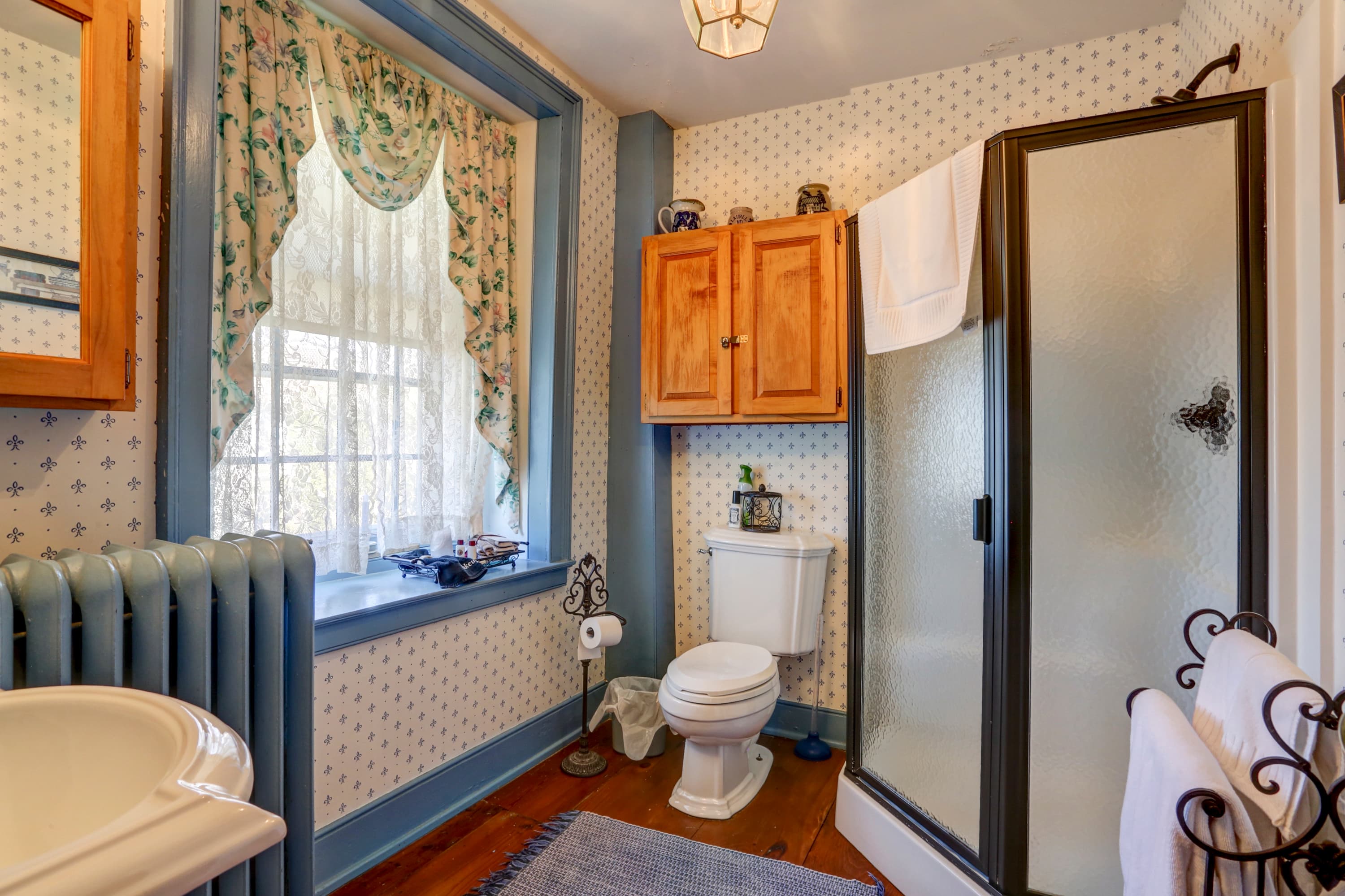 A bright and airy bathroom with a shower, a sink, a toilet, a window with a curtain, and a patterned rug on the floor.