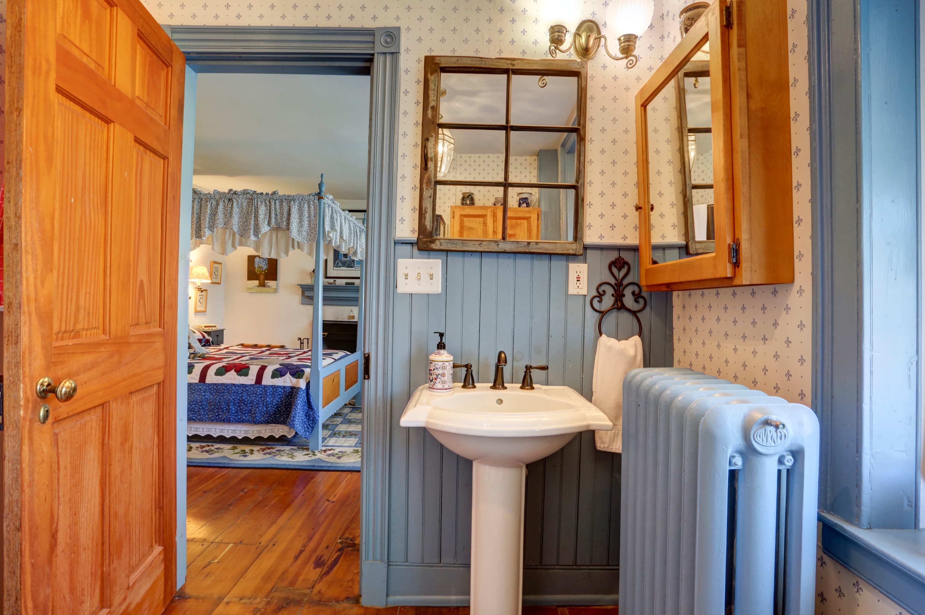 A small bathroom with a pedestal sink, a toilet, a radiator, and a view of a bedroom with a bed, a window, and blue walls through an open door.