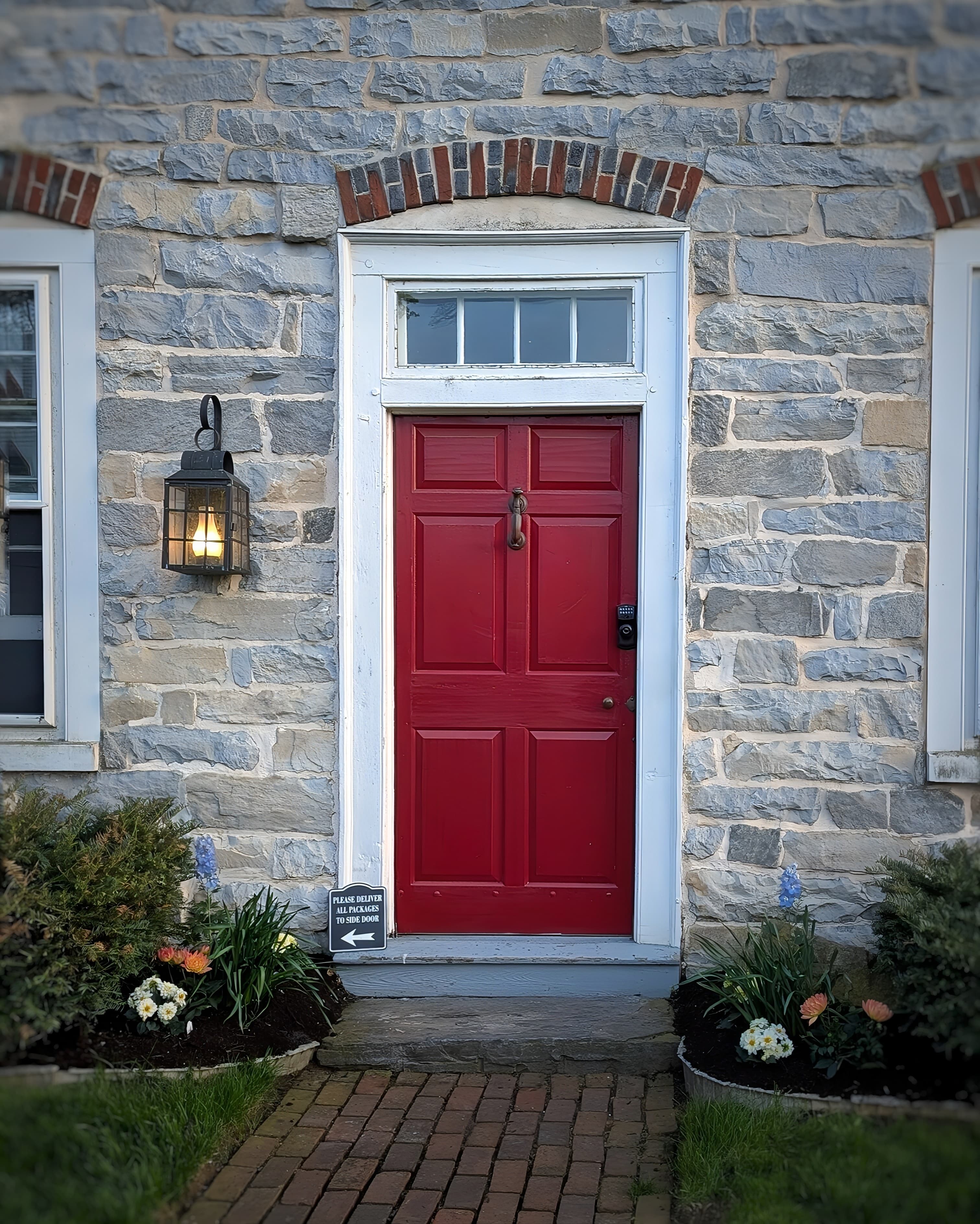 A red door on a stone house, flanked by flowers and a lantern, with a sign that reads "Please use side door." A red door on a stone house, flanked by flowers and a lantern, with a sign that reads "Please use side door."